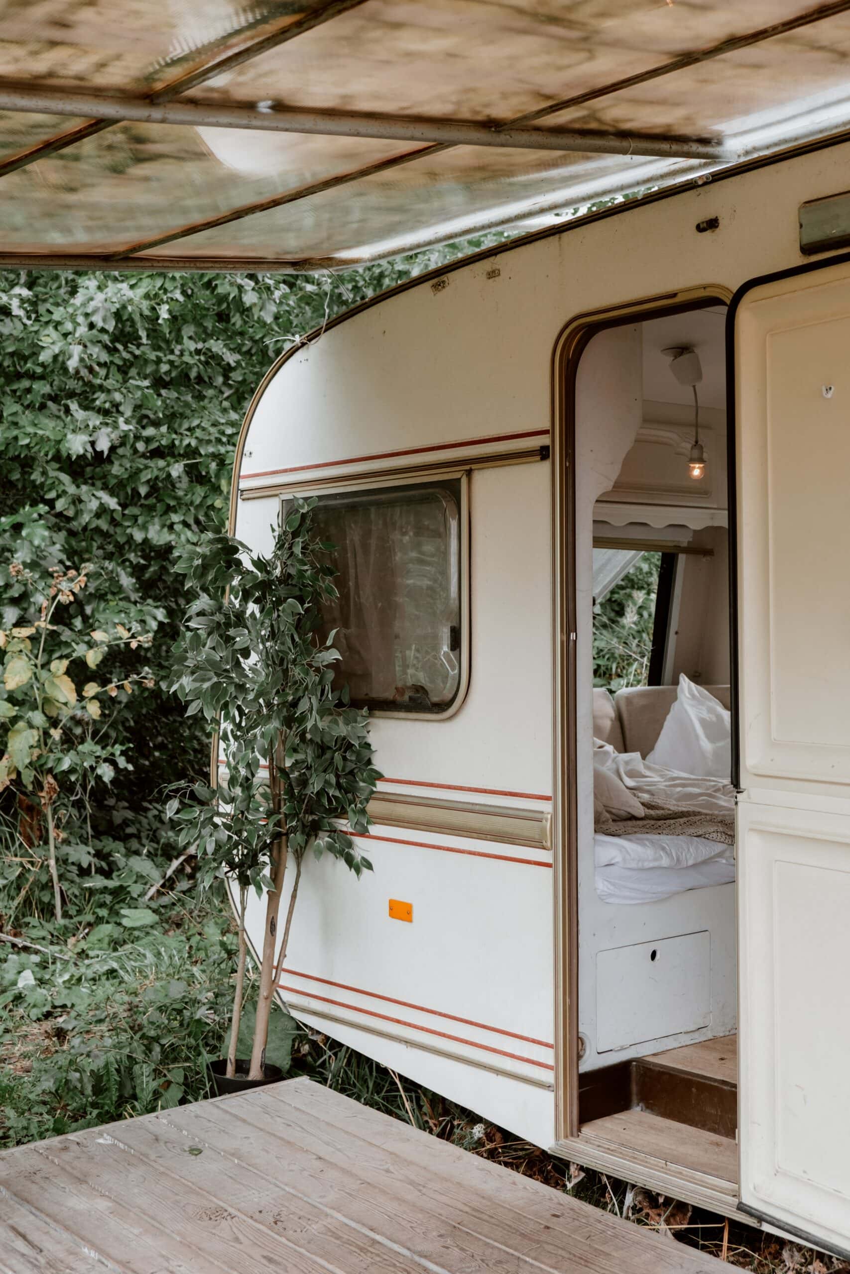 A white camper trailer with an open door, revealing a cozy interior with white bedding. It's parked on a wooden deck surrounded by lush greenery, with partial shade from an overhead covering.