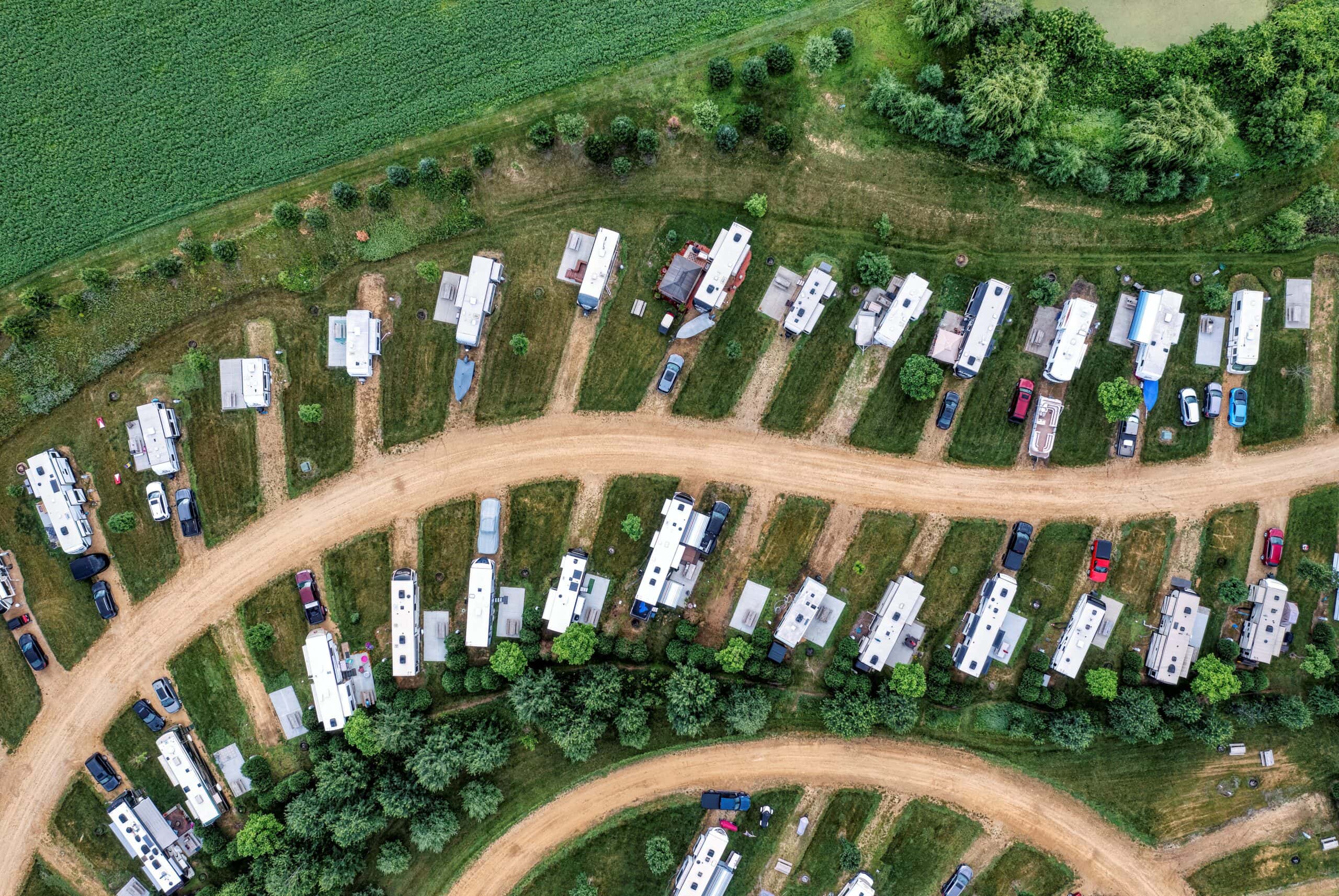 Aerial view of a campground with rows of RVs parked on grassy lots. Dirt roads wind through the area, surrounded by green trees and adjacent to farmland.