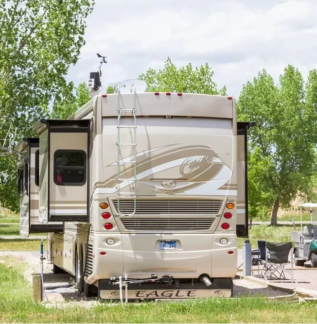 A large beige RV parked at a campsite with trees in the background. The RV has an extended slide-out and a ladder on the back. Outdoor chairs and a small table are set up to the right. A camper van is visible nearby.