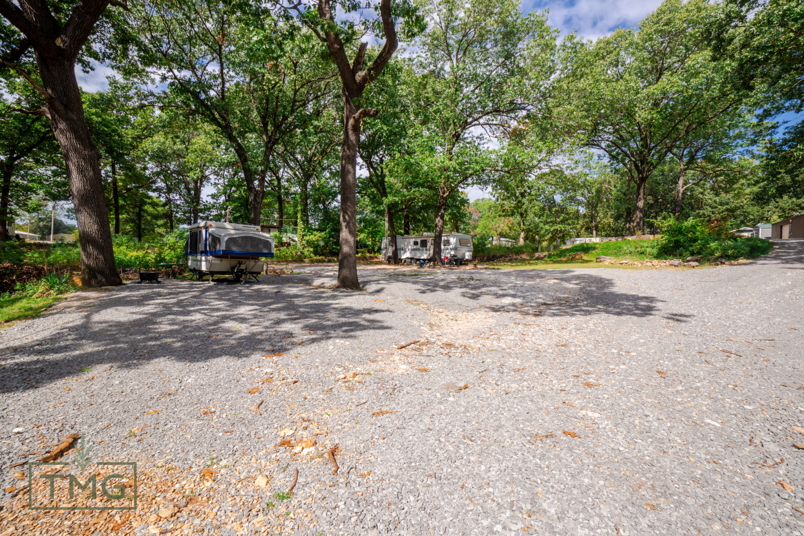 A gravel parking area with two RVs parked among large trees, surrounded by greenery under a partly cloudy sky.