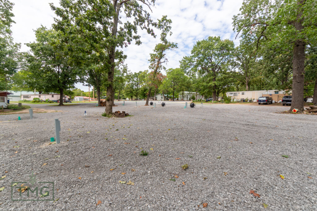 A gravel lot with scattered trees and utility hookups, surrounded by parked vehicles and mobile homes under a partly cloudy sky.