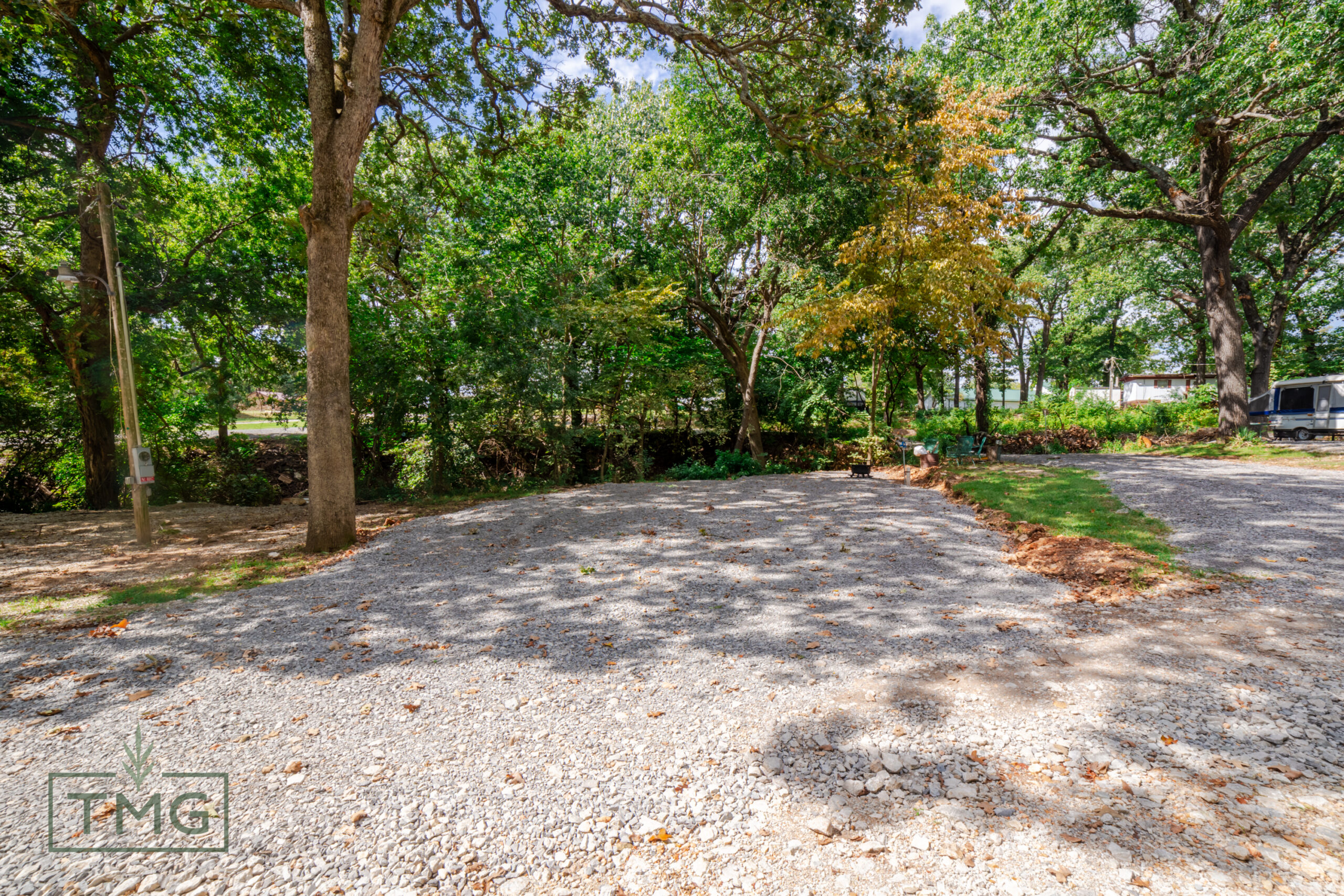 A gravel parking pad surrounded by trees and greenery on a partly sunny day, with a TMG logo in the bottom left corner.
