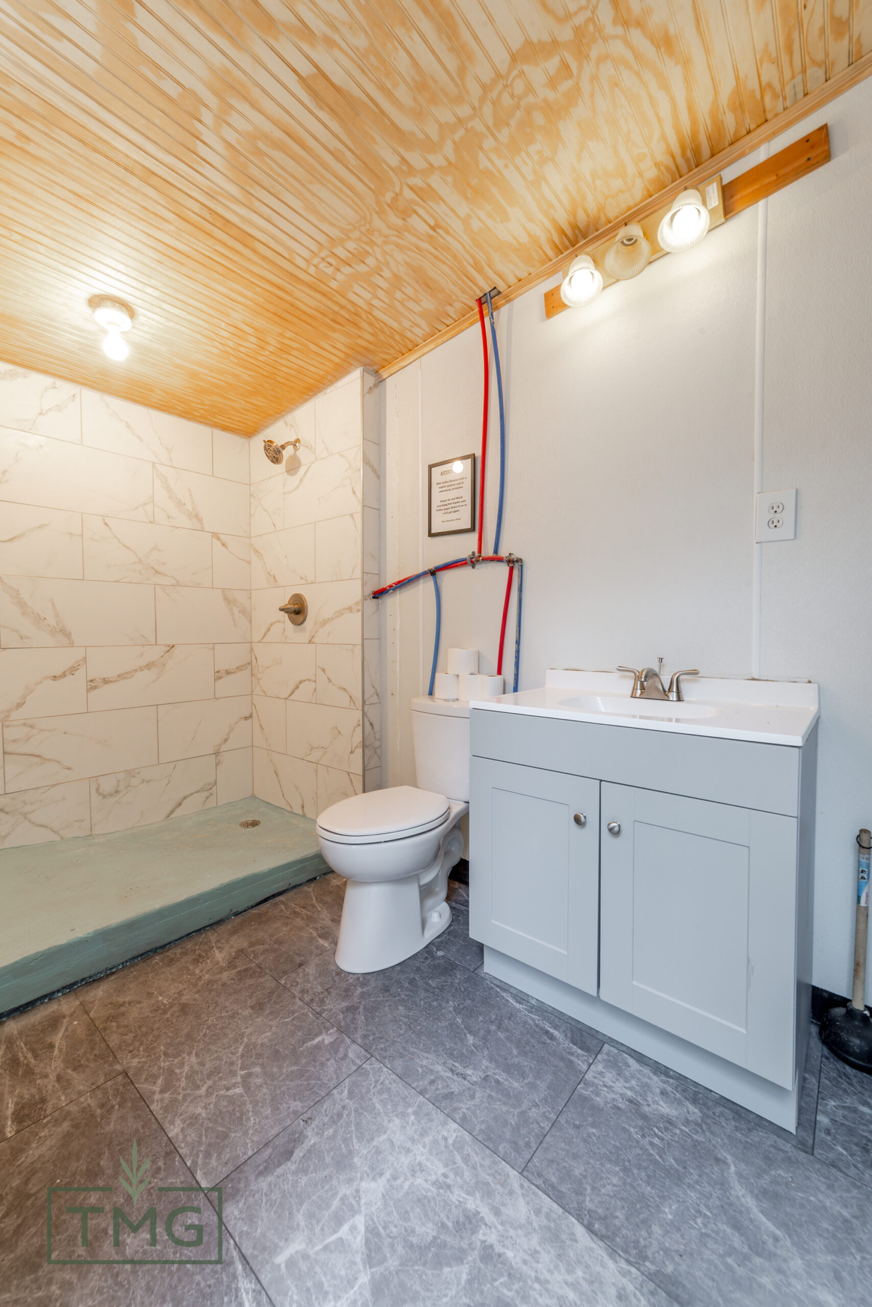 Bathroom with marble-look tile shower, gray vanity with sink, toilet, exposed plumbing, and wood-paneled ceiling.